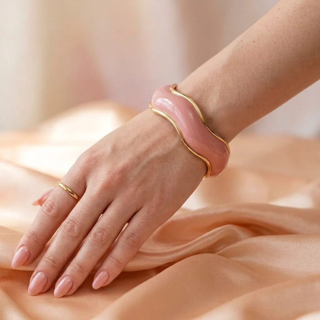 MAYA 01 Saint-Tropez wave cuff in Petal Pink worn on the wrist with a fine gold ring, on peach satin