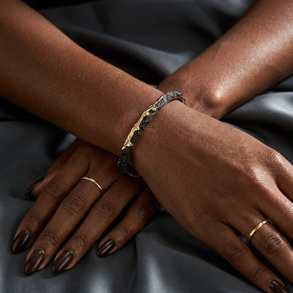 MAYA 01 Santorini cuff in Volcanic Gold worn on the wrist with fine gold rings, resting on charcoal satin